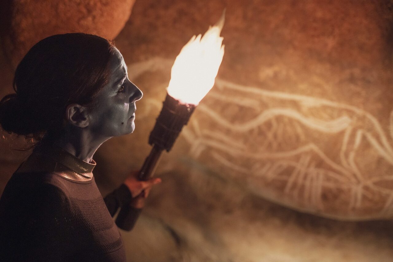 Pièce de théâtre L'œil de Dieu - Une femme, le visage maquillé de bleu, tient une torche qui éclaire les dessins d'une paroi de la grotte restituée.