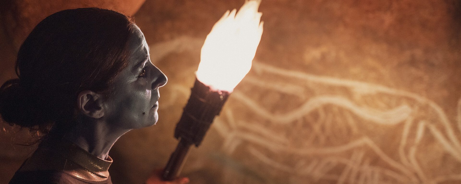 Une femme, le visage maquillé de bleu, tient une torche qui éclaire les dessins d'une paroi de la grotte restituée.