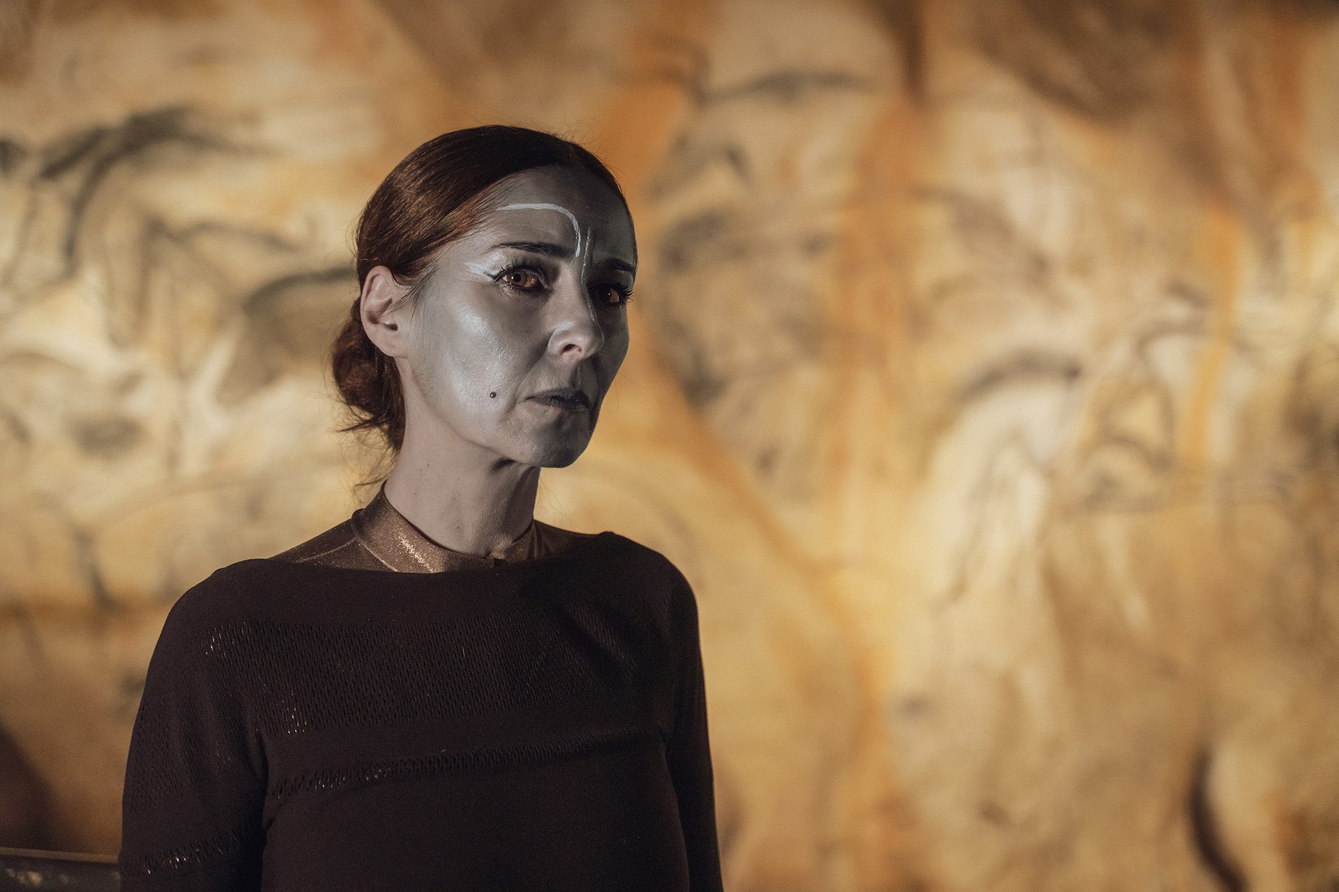 Pièce de théâtre L'œil de Dieu - Une femme le visage maquillé de bleu nous regarde. Derrière elle, les peintures de la grotte Chauvet restituée.